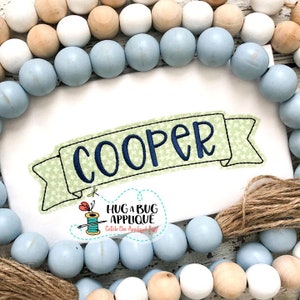 Könnte beinhalten: Gestickte Banner mit dem Namen "COOPER" in blauer Schrift. Das Banner ist grün mit einem weißen Sternenmuster und hat einen weißen Rand. Das Banner ist von einer Girlande aus blauen und beigefarbenen Holzperlen umgeben.
