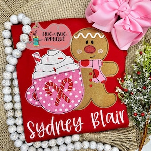 May include: Red Christmas-themed t-shirt with an embroidered gingerbread man holding a pink and white mug with whipped cream and candy canes. The text "Hug a Bug Applique" and "Sydney Blair" are also embroidered on the shirt.