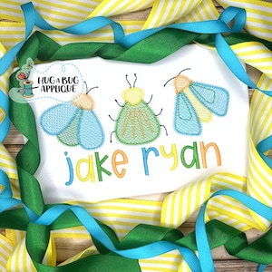 Op de afbeelding: Een witte romper met geborduurde blauwe, groene en gele vuurvliegjes en de namen "jake ryan" in kleurrijke letters.