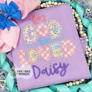 May include: A purple shirt with the text "for God so loved" in colorful patchwork letters. The name "Daisy" is embroidered in purple below the text. The shirt is surrounded by a decorative border of white beads and turquoise ribbon.