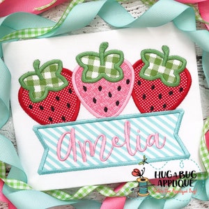 May include: Embroidered applique design featuring three red and pink strawberries with green leaves and a banner with the name Amelia in pink cursive lettering.