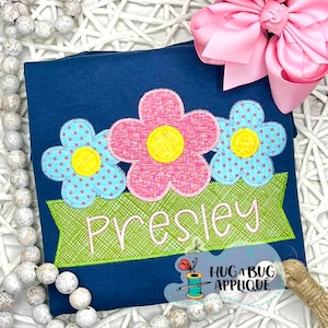 May include: A navy blue t-shirt with a green banner that says "Presley" and three appliqued flowers. The flowers are pink, blue, and yellow with white polka dots.