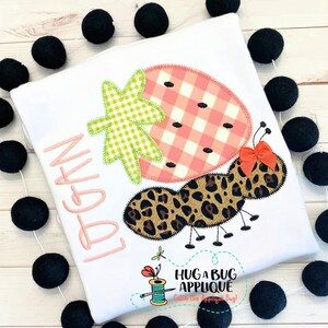 May include: A white t-shirt with the name "Logan" embroidered in pink. The shirt features an applique of a strawberry with a green leaf and a leopard print ant with a red bow.