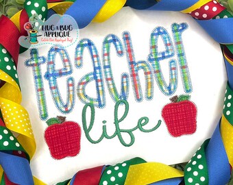 May include: White shirt with the text "teacher life" in colorful plaid and green script. Two red embroidered apples are on either side of the text.