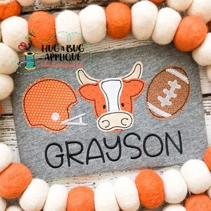 Puede incluir: Una camiseta gris con un casco de fútbol naranja, una pelota de fútbol marrón y una cabeza de vaca marrón y blanca con el nombre "GRAYSON" bordado en negro.