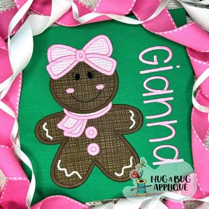 May include: A green shirt with a gingerbread person design, including a pink bow, scarf, and buttons. The name "Gianna" is embroidered vertically. The design includes the text "HUG A BUG APPLIQUE".