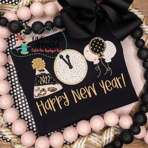 May include: Black t-shirt with gold embroidered "Happy New Year!" text, a party hat with "2022", a clock, and balloons. The shirt is decorated with gold and pink accents. A black bow and beaded garland surround the shirt.