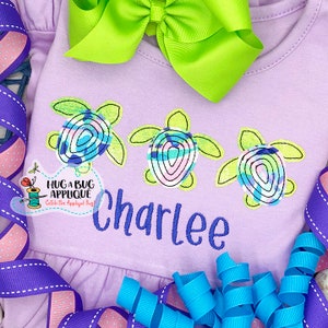 May include: A lavender-colored baby onesie with three embroidered sea turtles in shades of green, blue, and pink. The onesie is personalized with the name "Charlee" in blue embroidery.