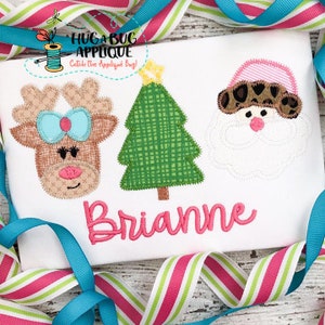 May include: A white shirt with a pink, green, and brown applique design of a reindeer, a Christmas tree, and Santa Claus. The name "Brianne" is embroidered in pink below the design.