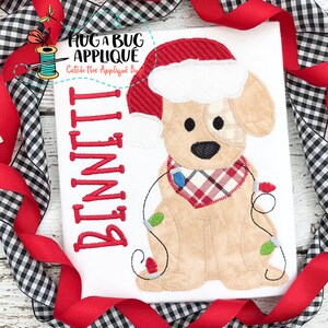 May include: A white t-shirt with a red embroidered applique of a dog wearing a Santa hat and a red and white plaid scarf. The dog is holding a string of Christmas lights. The text "BENNETT" is embroidered in red on the left side of the shirt.