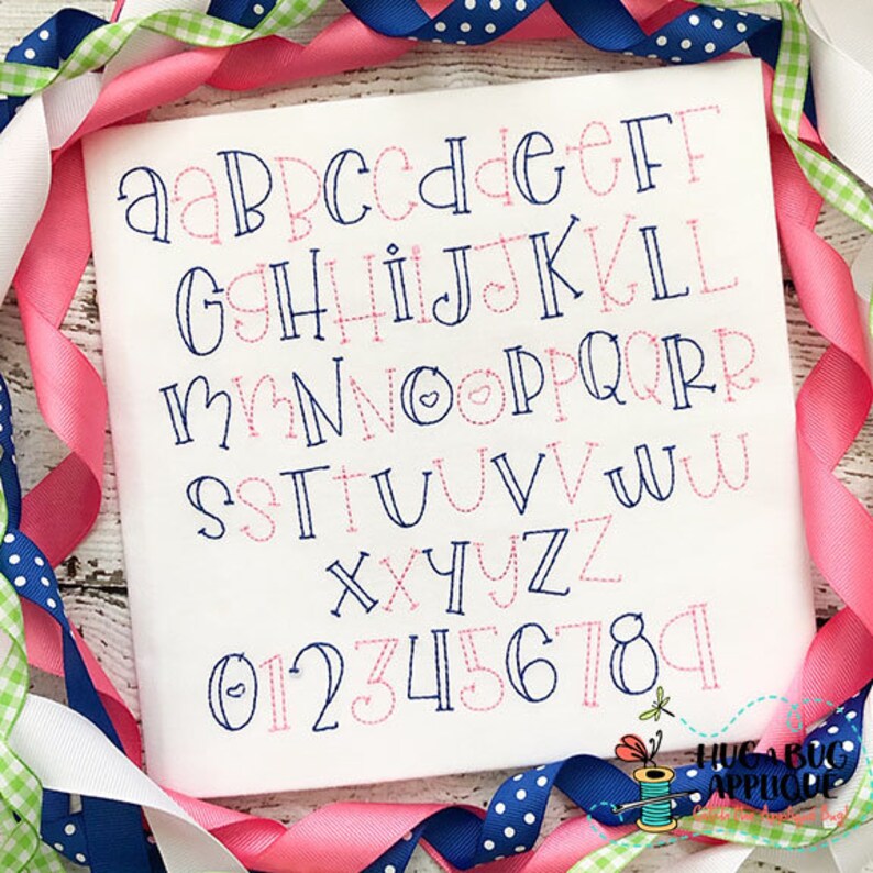 May include: A white fabric with a blue and pink alphabet and numbers embroidered on it. The letters are in a whimsical font with some letters having a heart shape in the middle. The fabric is surrounded by pink and blue ribbon with white polka dots.