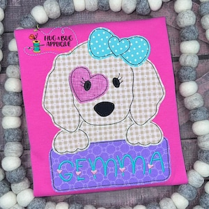 May include: An embroidered applique of a dog with a blue bow and a pink heart over one eye. The dog is holding a purple sign with the name "GEMMA" embroidered in turquoise.