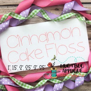 Pode incluir: Desenho bordado do texto "Cinnamon Cake Floss" em fio rosa sobre um fundo de tecido branco. O design está disponível nos tamanhos 2,5 cm, 3,8 cm, 5 cm, 6,3 cm, 7,6 cm e 8,9 cm.