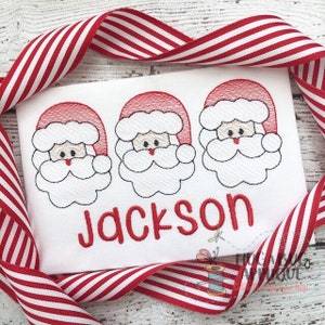 Op de afbeelding: Wit geborduurd shirt met drie rode kerstmangezichten en de naam "Jackson" in rode letters.