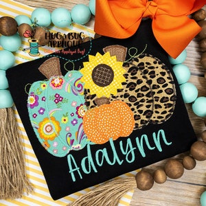 Op de afbeelding: Zwart T-shirt met een kleurrijk appliqué-ontwerp van drie pompoenen en een zonnebloem. De pompoenen zijn versierd met bloemen-, luipaardprint- en stippenpatronen. De naam "Adalynn" is in turquoise op het shirt geborduurd.