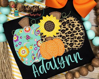May include: Black t-shirt with a colorful appliqué design of three pumpkins and a sunflower. The pumpkins are decorated with floral, leopard print, and polka dot patterns. The name "Adalynn" is embroidered in teal on the shirt.
