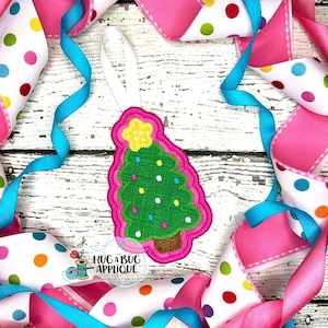 Puede incluir: Un adorno de árbol de Navidad de fieltro rosa con un árbol de fieltro verde, estrellas de fieltro blanco y una estrella de fieltro amarilla en la parte superior. El adorno tiene un lazo para colgar.
