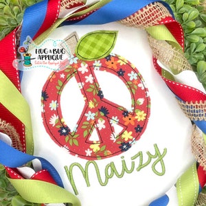 May include: A white fabric applique with a red peace sign with a floral pattern. The peace sign is inside a red apple shape with a green leaf. The name "Maizy" is embroidered in green below the apple.