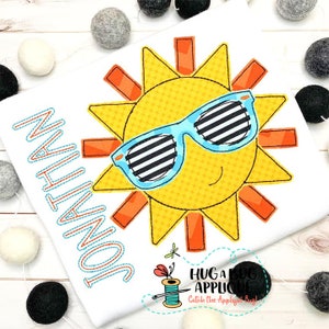 May include: A yellow sun with a smiling face wearing blue sunglasses with black stripes. The sun has orange rays and is on a white background with the name "JONATHAN" stitched in blue and orange thread.