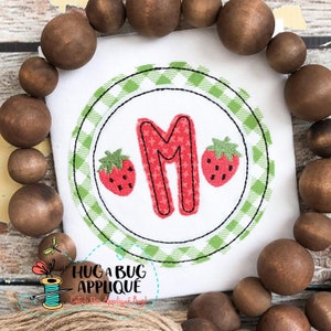 May include: A white embroidered applique with a green and white checkered circle. The circle contains a red letter M with a white star pattern and two red strawberries with green leaves.