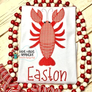 May include: A white t-shirt featuring a red and white gingham lobster applique and the name Easton embroidered in red underneath. The shirt is surrounded by a red and white beaded garland.