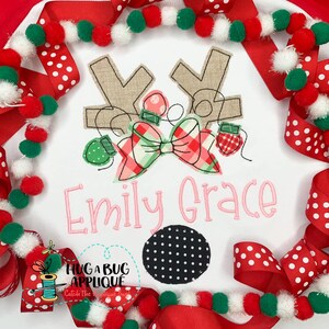 May include: A white onesie with a brown reindeer applique with a red and green plaid bow and Christmas lights. The onesie is personalized with the name "Emily Grace" in pink lettering. The onesie is surrounded by red and white polka dot ribbon and a garland of white, red, and green pom-poms.