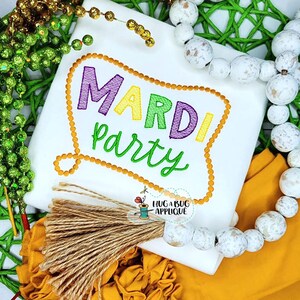 May include: White shirt with embroidered text that says "Mardi Party" in a colorful banner with a gold tassel. The shirt is surrounded by beads and a wooden bead necklace.