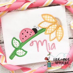 Puede incluir: Una camiseta blanca con un tallo verde, una mariquita rosa y una aplicación de girasol amarillo. El nombre "Mia" está bordado en letras cursivas rosas.