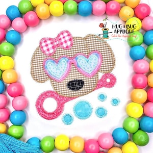 Puede incluir: Diseño de aplique de un perro marrón que lleva gafas de sol en forma de corazón rosa y un lazo rosa. El perro sostiene una varita de burbujas rosa y hay burbujas azules a su alrededor. El diseño está sobre un fondo de tela blanca.