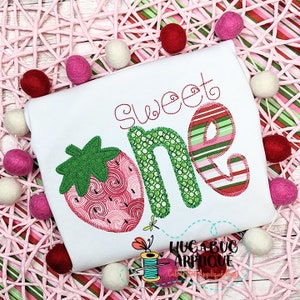May include: White t-shirt with a strawberry and the words "Sweet One" in red and green embroidery. The strawberry has a red and pink swirl design. The letter "O" has a striped pattern. A cute design for a baby's first birthday.