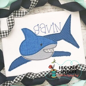 May include: A blue and white shark applique with the name "gavin" stitched above it. The applique is on a white fabric background.