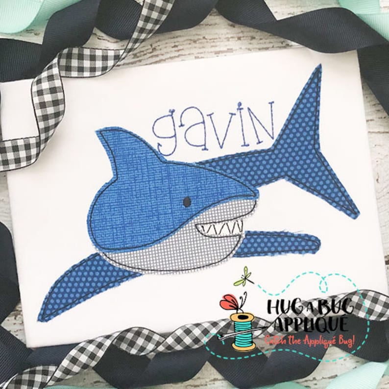 May include: A blue and white shark applique with the name "gavin" stitched above it. The applique is on a white fabric background.