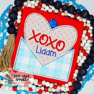 Può includere: Maglietta rossa con un design a busta a quadri blu chiaro e bianco. La busta ha un cuore beige con pois rossi e il testo "xoxo Liam" in rosso.