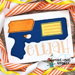 May include: A blue and orange embroidered design of a toy gun with the name Elijah stitched underneath. The design is on a white fabric background.