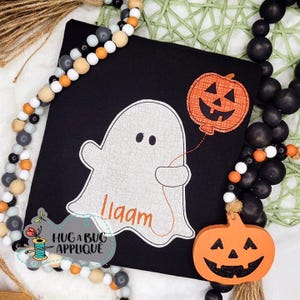 Puede incluir: Cuadrado negro con un fantasma blanco sosteniendo un globo de calabaza naranja. El fantasma tiene el nombre "liaam" en naranja. Una calabaza de madera y una guirnalda de cuentas rodean el cuadrado. El texto "HUG A BUG APPLIQUE" también es visible.