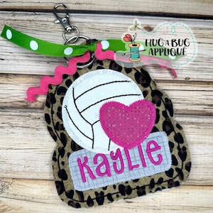 May include: A brown and black leopard print fabric keychain with a white volleyball, a pink heart, and the name "kaylie" embroidered in pink.