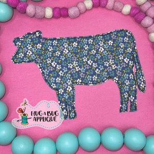 May include: A blue cow applique with a white floral pattern. The applique is on a pink background with a turquoise bead garland around the edge. The text "Hug a Bug Applique" is in the lower left corner.
