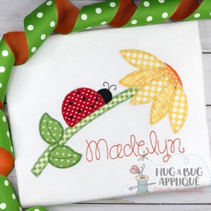 May include: A red and black ladybug applique with white polka dots on a green stem with leaves. The stem is attached to a yellow flower with white polka dots. The name "Madelyn" is embroidered in red below the applique. The text "Hug a Bug Applique" is also embroidered below the name.