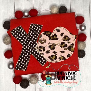 Puede incluir: Camiseta roja con una aplicación en forma de corazón marrón y blanco con lunares blancos y una aplicación en forma de labio de estampado de leopardo rosa y dorado. El texto "Hug a Bug Applique" y "Catch the Applique Bug!" está impreso en la camiseta.