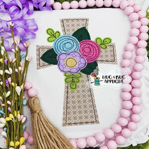 May include: Embroidered cross with a floral design. The cross is brown with a white outline and the flowers are pink, blue, and purple.