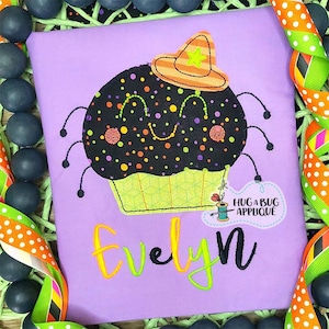 May include: Purple children's t-shirt with a black and orange cupcake with a smiling face and a hat. The cupcake has a green and white icing design. The text "Evelyn" is embroidered on the shirt. The text "Hug a Bug Applique" is also on the shirt.