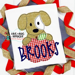 May include: A brown and white dog applique holding a red and white checkered apple with the name "BROOKS" embroidered in blue on the apple.