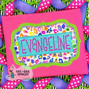 May include: A pink fabric item with the name "EVANGELINE" embroidered in pink on a turquoise rectangle. The rectangle is set within a green scalloped frame, surrounded by a leopard print design. The background is pink, with decorative ribbons.