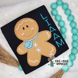 May include: A black fabric item featuring a gingerbread man applique. The gingerbread man is light brown with a blue scarf and white icing details. The name "LIAM" is embroidered in blue. The logo "HUG A BUG APPLIQUE" is also visible.