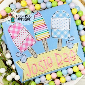 Puede incluir: Una pancarta azul y amarilla con el nombre "Josie Rae" bordado en ella. Tres paletas de helado bordadas están por encima de la pancarta, una con cuadros rosa y blanco, otra con rayas arcoíris y otra con cuadros azul y blanco.