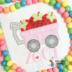 Puede incluir: Camiseta blanca con un dise&ntilde;o bordado de un vag&oacute;n rosa lleno de fresas rojas. El vag&oacute;n tiene ruedas negras y el nombre "Zoe" en escritura rosa. Un peque&ntilde;o coraz&oacute;n rojo est&aacute; al lado del nombre.