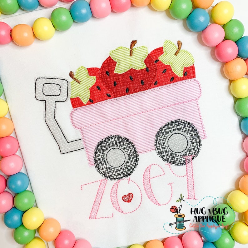 Puede incluir: Camiseta blanca con un dise&ntilde;o bordado de un vag&oacute;n rosa lleno de fresas rojas. El vag&oacute;n tiene ruedas negras y el nombre "Zoe" en escritura rosa. Un peque&ntilde;o coraz&oacute;n rojo est&aacute; al lado del nombre.