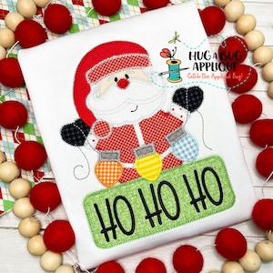 Puede incluir: Una camiseta blanca con una aplicación de Papá Noel sosteniendo una cadena de luces de colores. El texto "Ho Ho Ho" está en una pancarta verde debajo de Papá Noel.