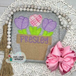May include: A gray shirt with a personalized flower pot applique. The pot has three flowers, one pink and two purple, with the name "PRESLEY" embroidered on the front. The shirt is decorated with a pink bow.
