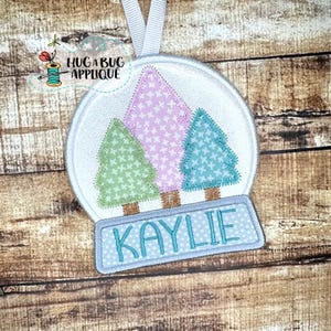 Puede incluir: Un adorno de bola de nieve blanca con tres árboles estilizados en su interior. Los árboles son rosa, verde y azul. La bola de nieve tiene una etiqueta de nombre personalizada azul y blanca que dice "KAYLIE".